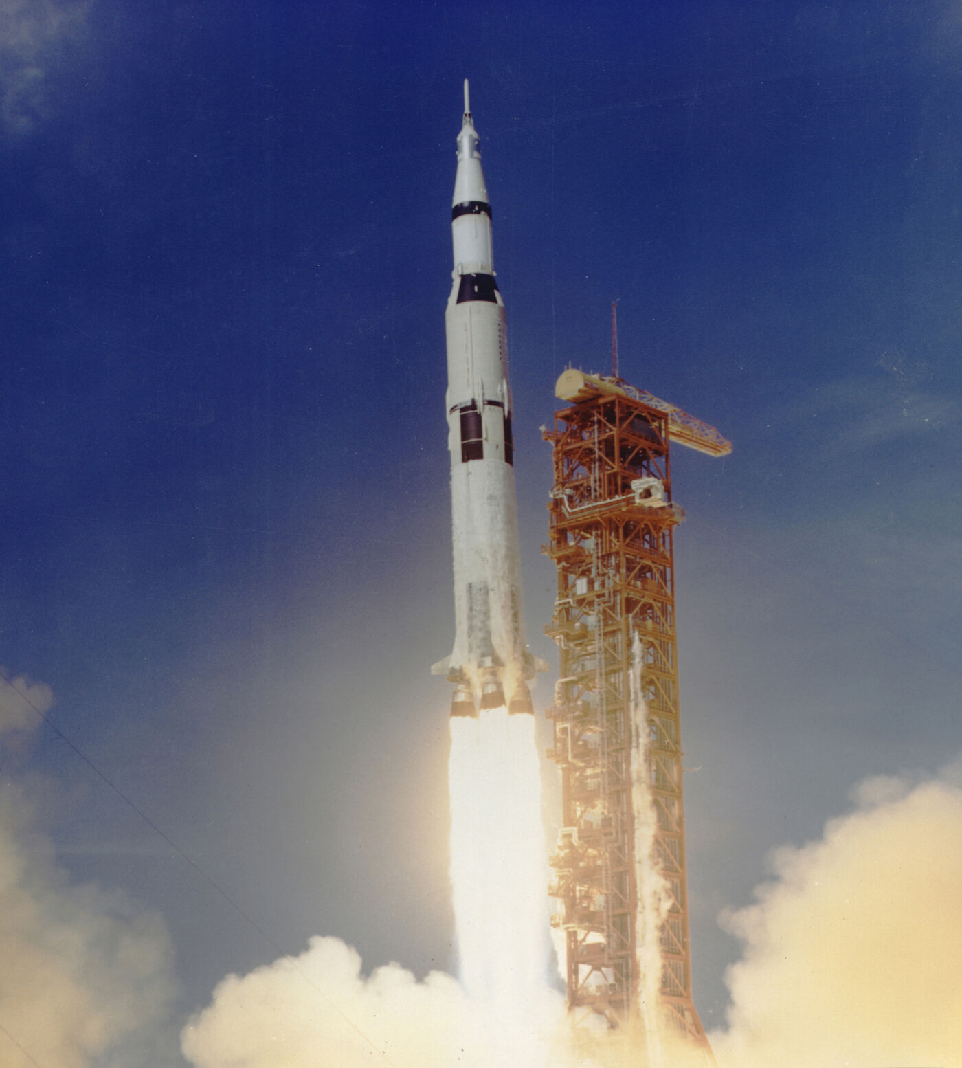 The Apollo 11 Saturn V rising past the launch tower at Kennedy Space Center on 16 July 1969, exhaust column glowing white at the base, blue sky above